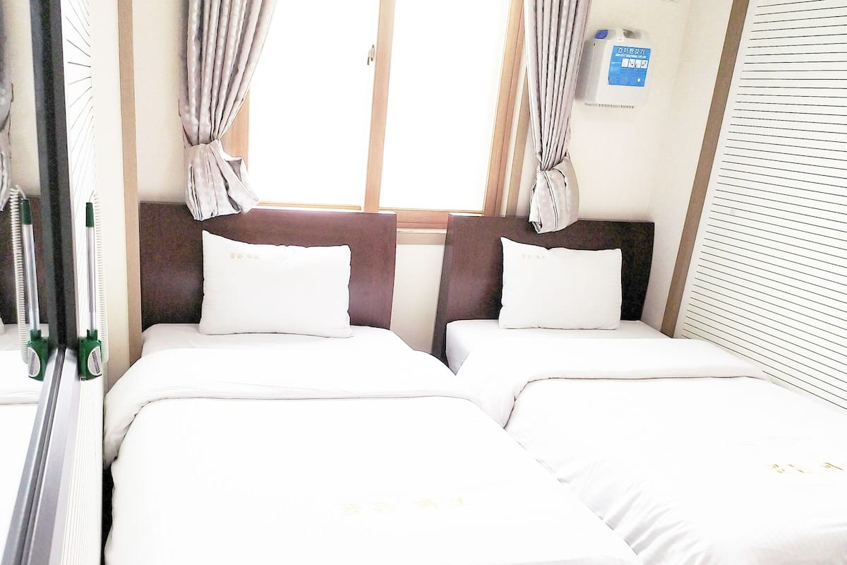 Une chambre d’hôtel avec deux lits, chacun doté de draps et d’oreillers blancs, d’une tête de lit en bois et d’une fenêtre avec des rideaux.
