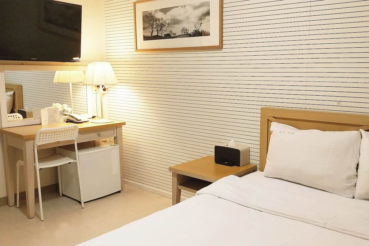 Une chambre d’hôtel avec un lit, un bureau avec une lampe, une chaise, une table de chevet avec un téléphone et un tableau encadré au mur.