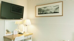 A hotel room with a flat-screen TV mounted on the wall, a desk with a telephone and a lamp, and a framed picture of a landscape.