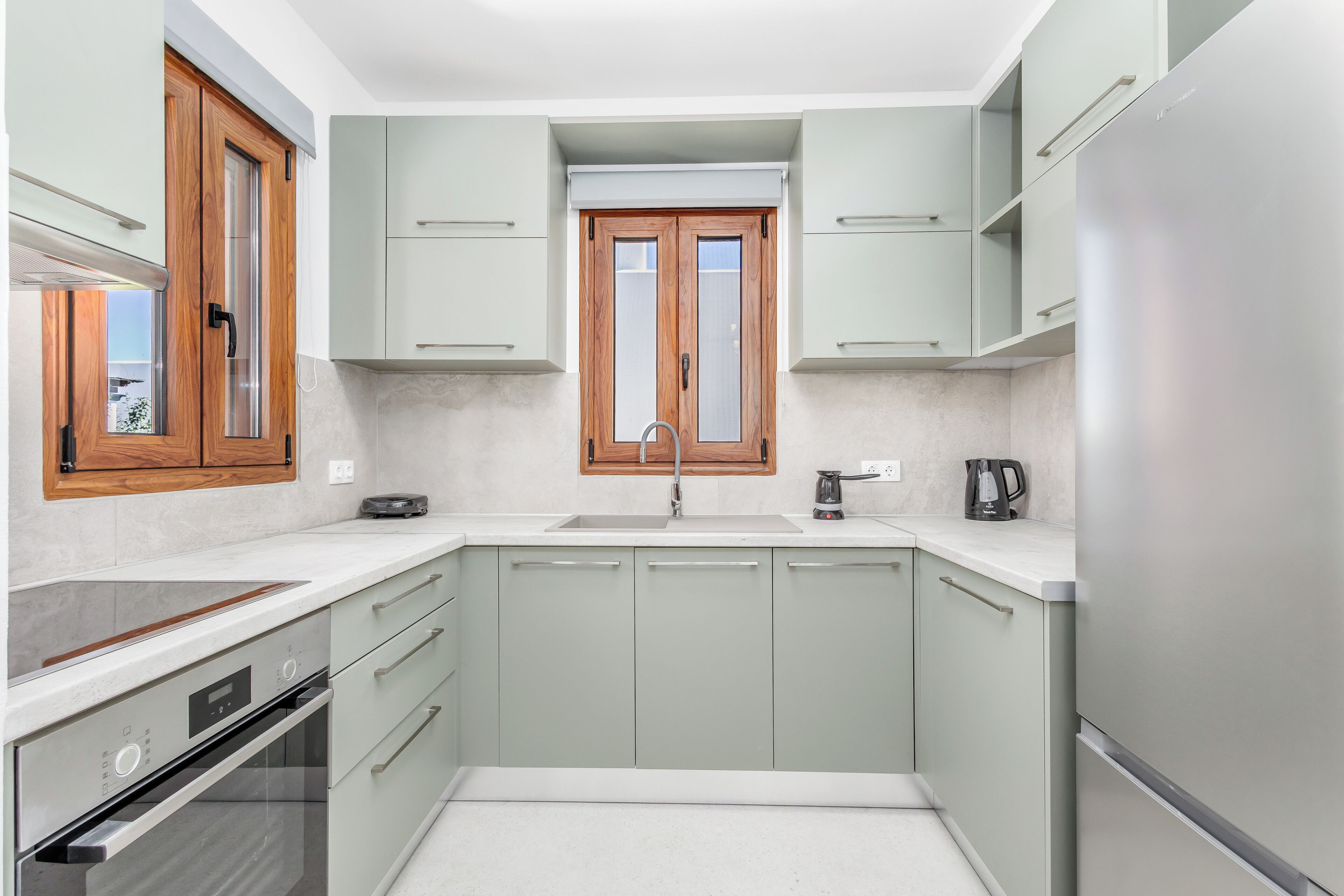 Een moderne keuken met lichtgroene kastjes, een roestvrijstalen koelkast, een inbouwoven en een raam boven de spoelbak.