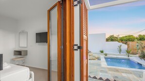 A modern room with a sliding glass door leading to a pool area, a flat-screen TV mounted on the wall, and a small seating area with a bench and table.