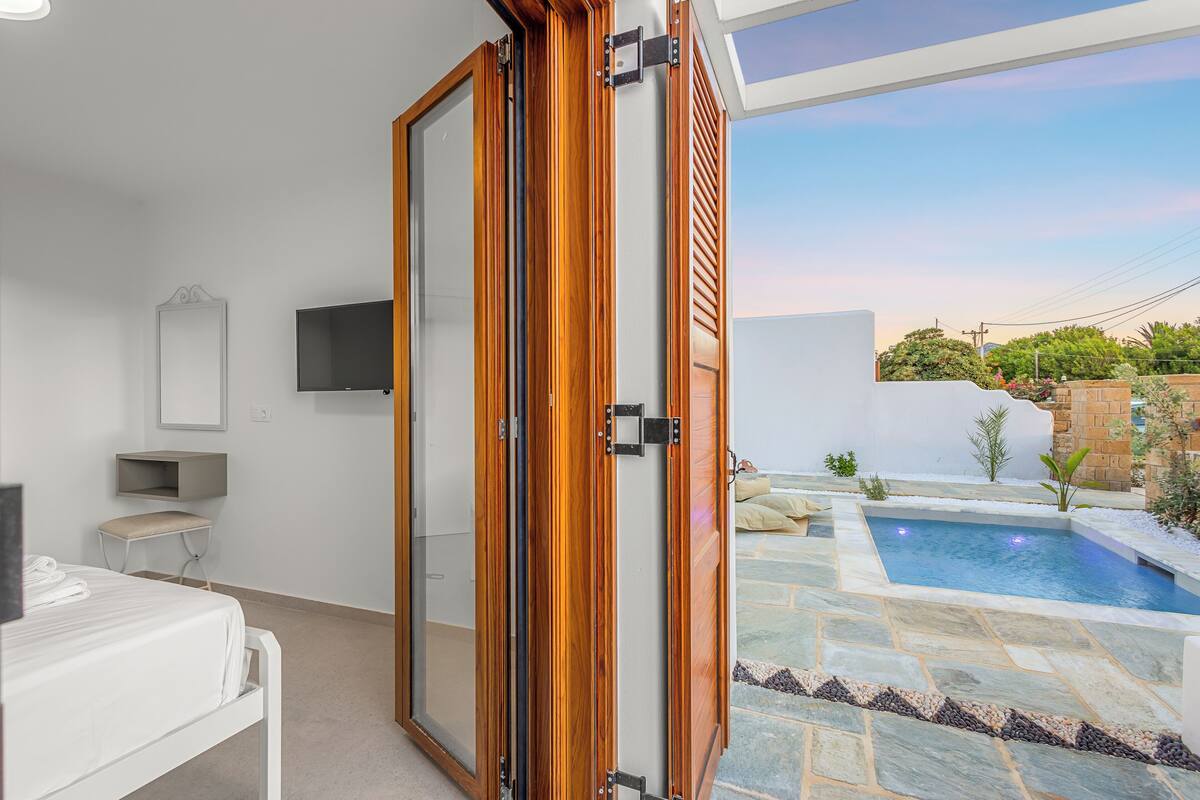 A modern room with a sliding glass door leading to a pool area, a flat-screen TV mounted on the wall, and a small seating area with a bench and table.