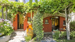 Ein Garteneingang mit einer Pergola, einer großen Vase und einem Backsteinhaus mit Tür und Fenstern.