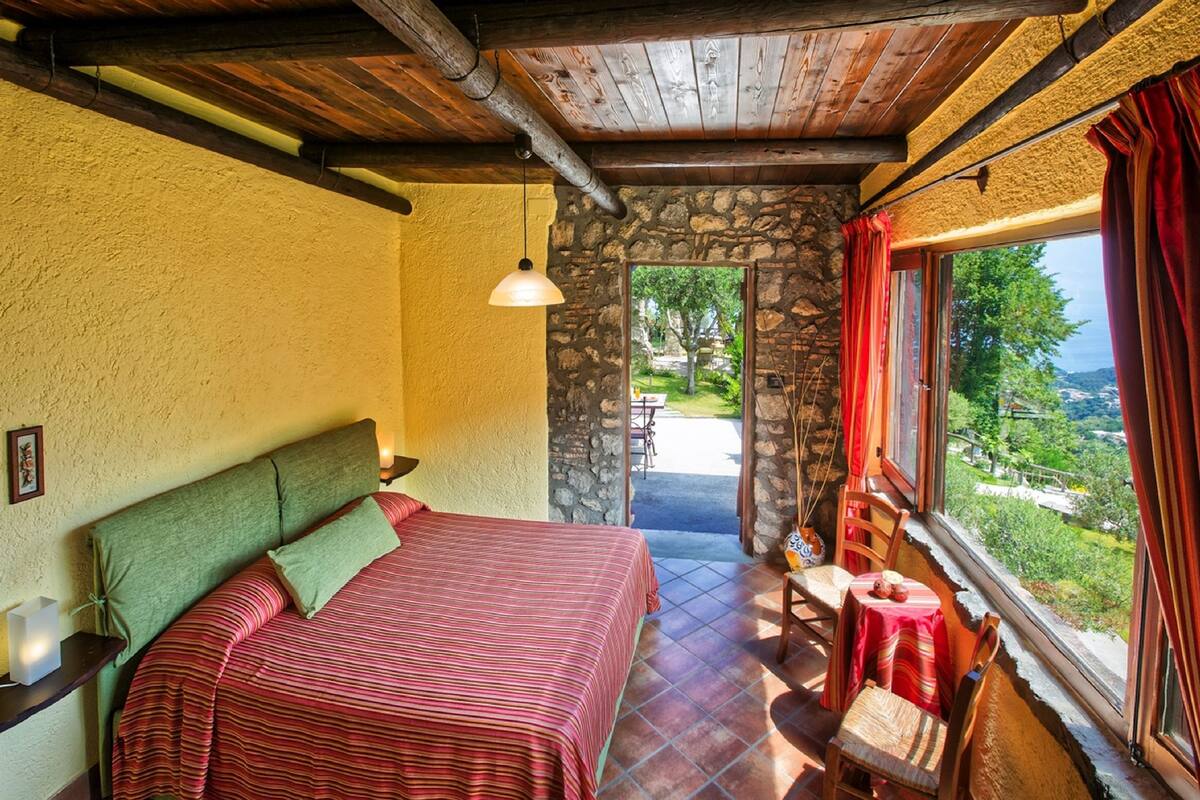 Une chambre à coucher comprenant un lit, une table de chevet, une table avec des chaises, une fenêtre avec des rideaux et une vue sur l’extérieur.