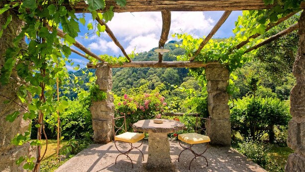 Ein Steintisch und zwei Stühle stehen unter einer mit Weinreben bewachsenen Pergola in einem Garten mit Blick auf einen bewaldeten Hang.