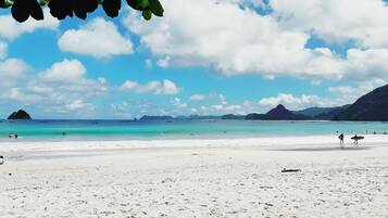 A beach with white sand, clear turquoise water, and distant mountains.