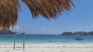 A beach with a thatched roof structure, a swing set, and a boat in the water. - Harmony Villas Lombok (Kuta)