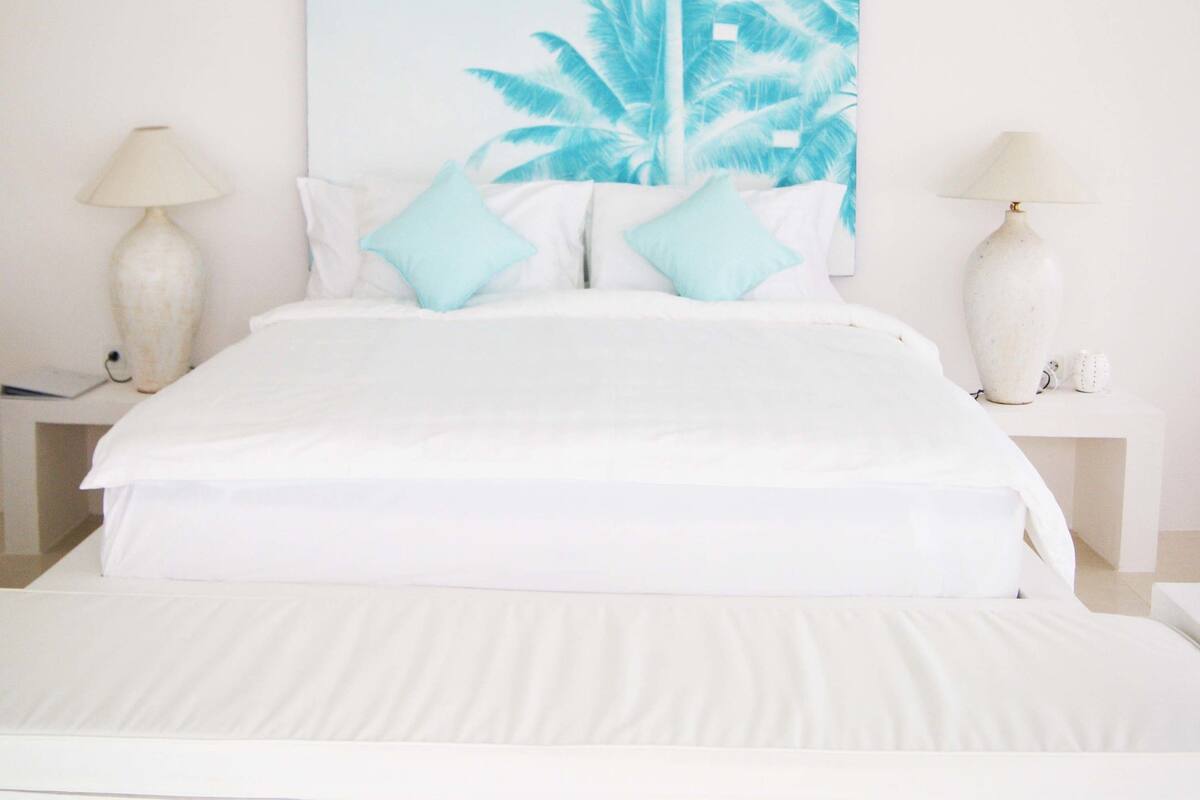 Un dormitorio con una cama grande, ropa de cama blanca, dos mesitas de noche con lámparas y un cuadro grande de una palmera tropical en la pared.