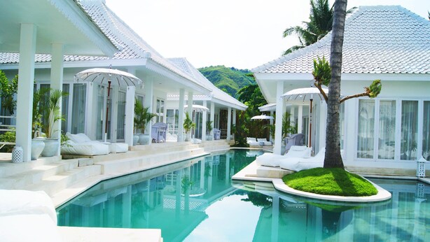 A pool area with lounge chairs and umbrellas, surrounded by white buildings with tiled roofs. - Harmony Villas Lombok (Kuta)