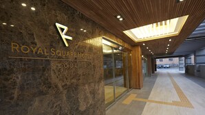 Exterior del Hotel Royal Square, con fachada de mármol, entrada de cristal y techo de madera con iluminación empotrada.