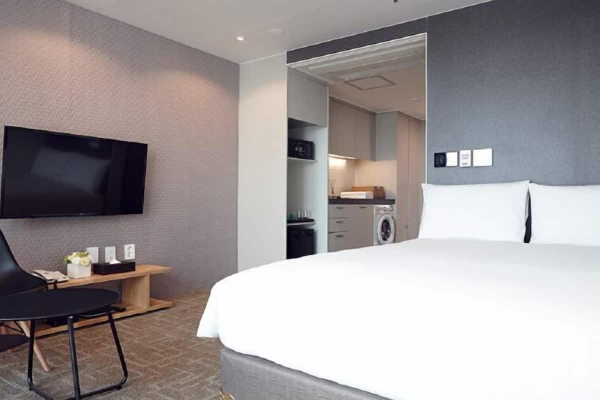 Una habitación de hotel moderna con una cama grande, un televisor de pantalla plana montado en la pared, un escritorio pequeño con una silla y una kitchenette visible.