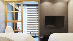 Habitación de hotel con dos camas, un televisor montado en la pared, una mesita pequeña y vistas a un balcón con persianas a través de una puerta.