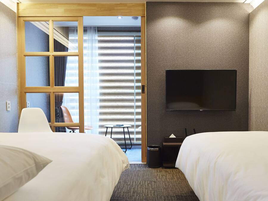A hotel room with two beds, a TV mounted on the wall, a small table, and a view through a door to a balcony with blinds.