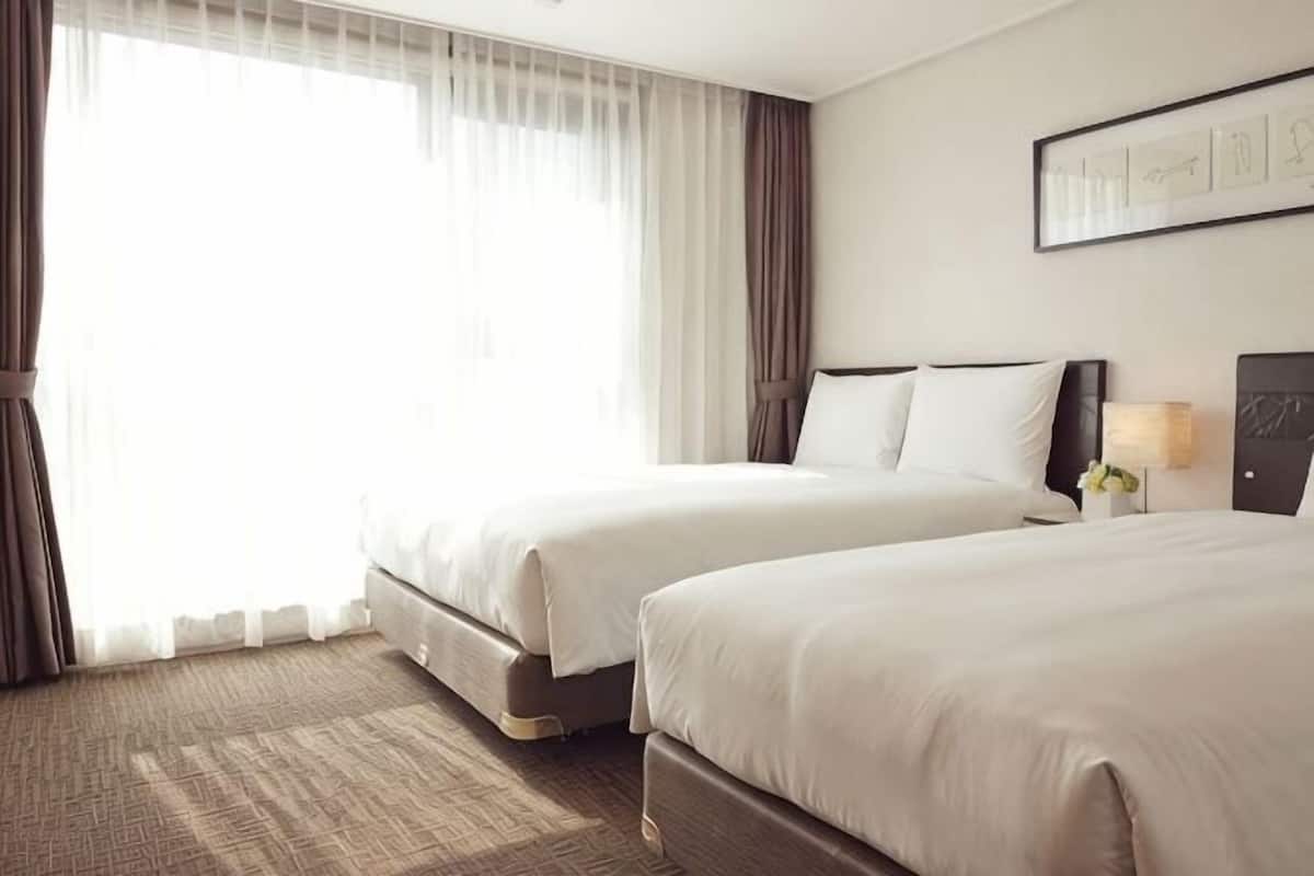 Habitación de hotel con dos camas, un ventanal con cortinas, un cuadro en la pared y una mesita de noche con lámpara.