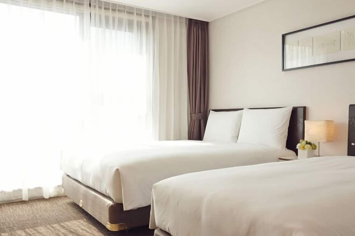 A hotel room with two beds, a large window with sheer curtains, a framed picture on the wall, and a bedside table with a lamp and flowers.