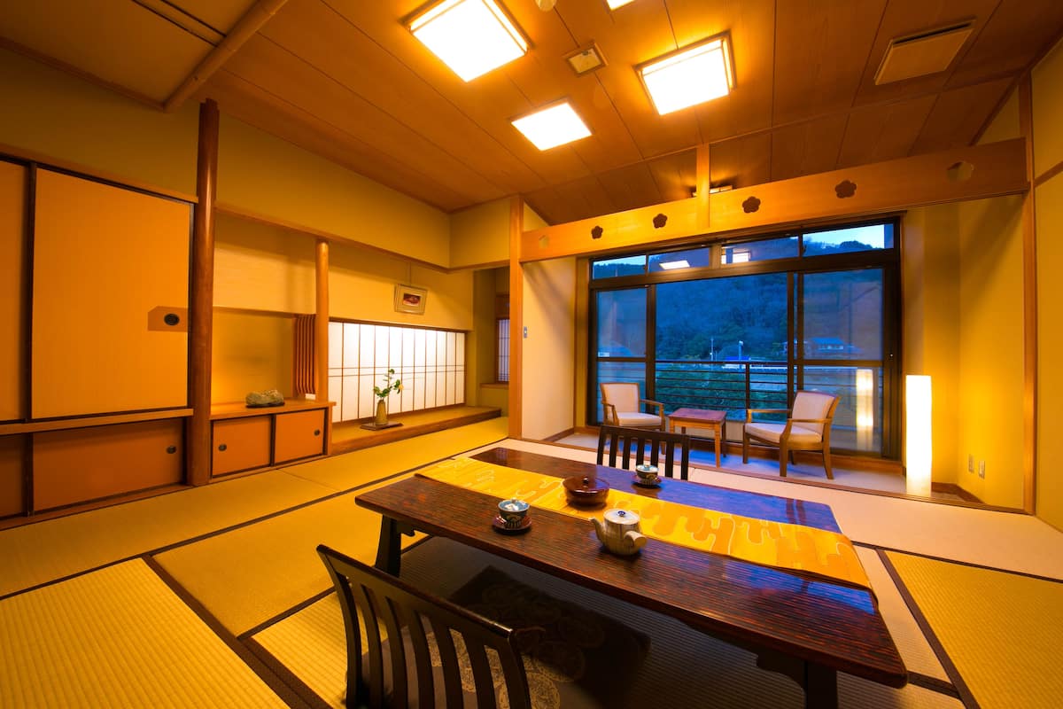 A traditional Japanese room with tatami flooring, a low table with a tea set, and a view of the outdoors through large windows.
