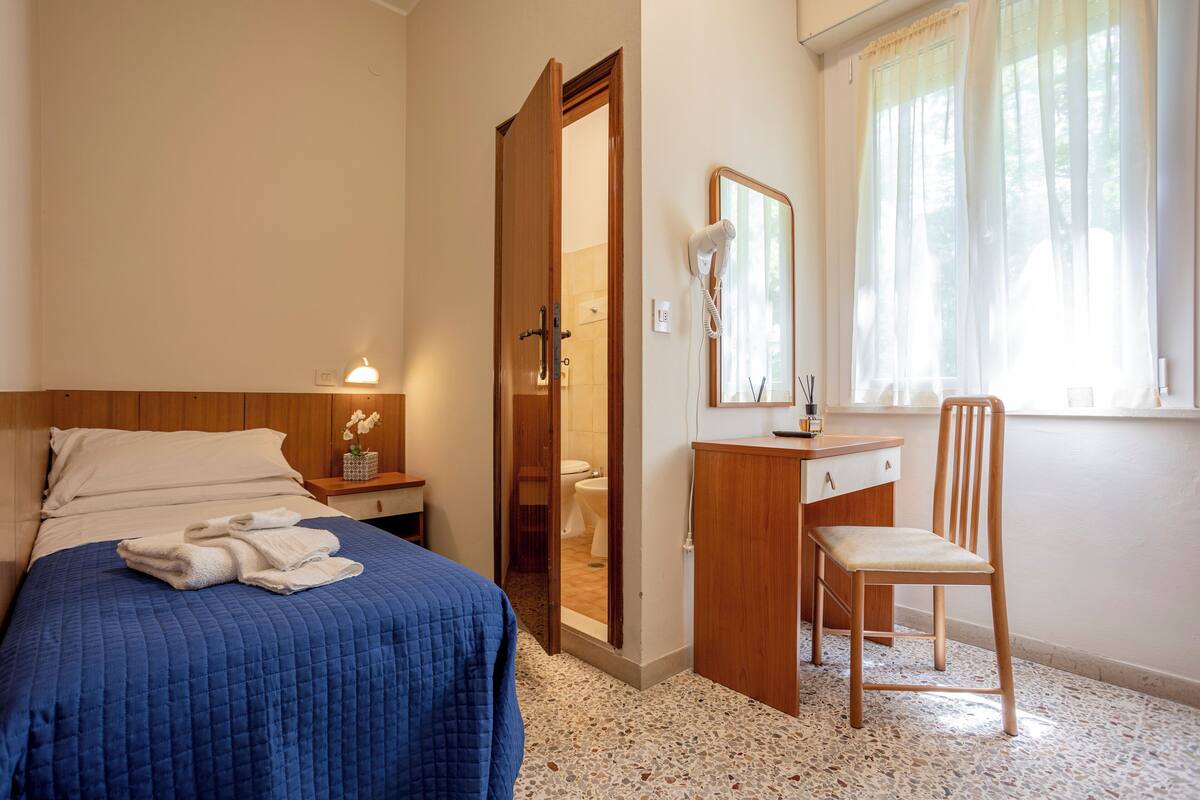 Un dormitorio con una cama, una mesita de noche de madera, un escritorio con una silla, un espejo y una ventana con cortinas.