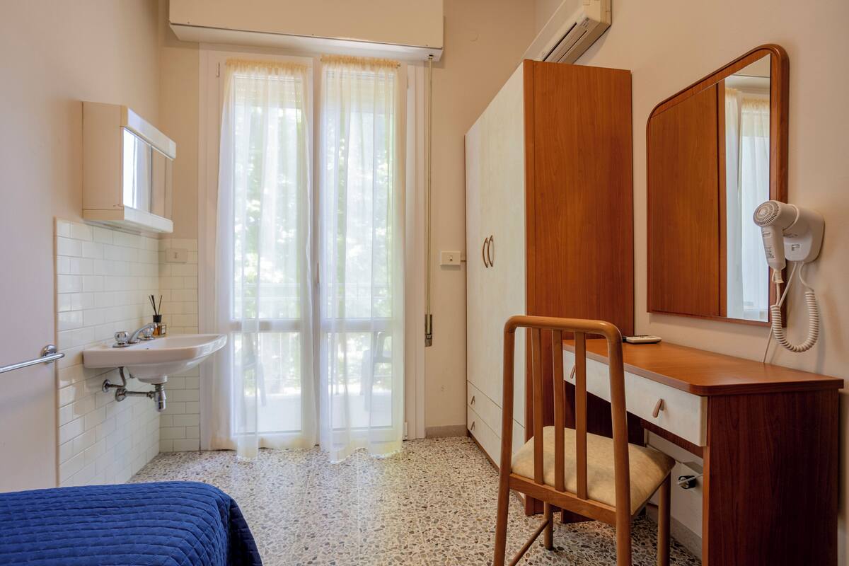 Un dormitorio con una cama, una silla de madera, un escritorio con espejo, un secador de pelo y una ventana con cortinas.