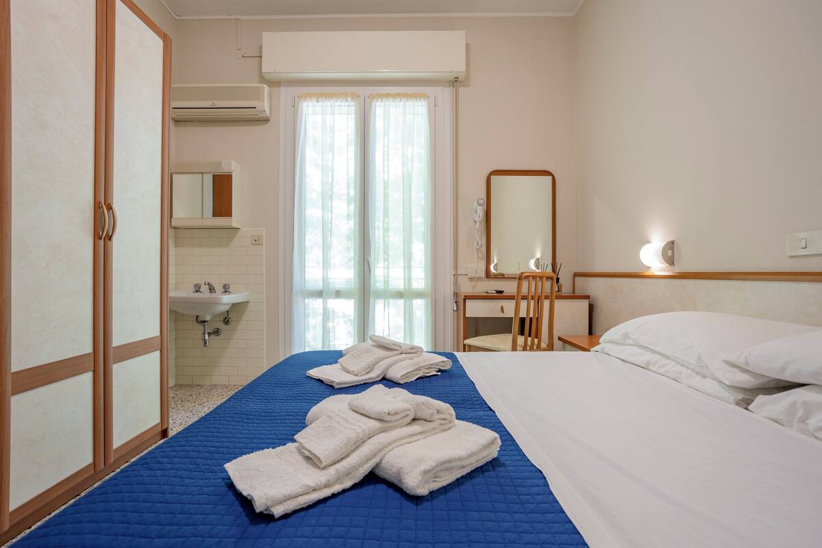 Habitación de hotel con cama, escritorio, silla, espejo y baño con lavamanos e inodoro.