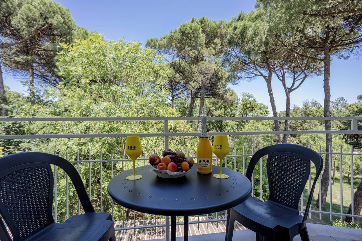 Un balcón con dos sillas, una mesa con frutero y dos botellas, y vista a árboles.