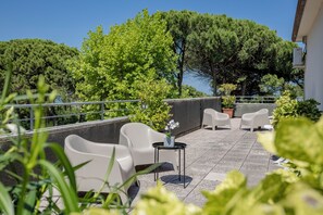 Una terraza con sillas blancas y una mesa pequeña, rodeada de plantas en macetas y árboles.