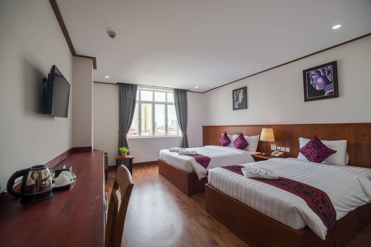 A hotel room with two beds, a wooden desk with a kettle, a TV, and a window with curtains.