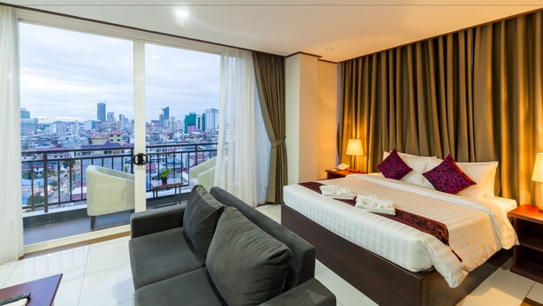 A hotel room with a large bed, a sofa, a balcony with a city view, and a cityscape visible through the windows. - Le Mont Hotel (Phnom Penh)