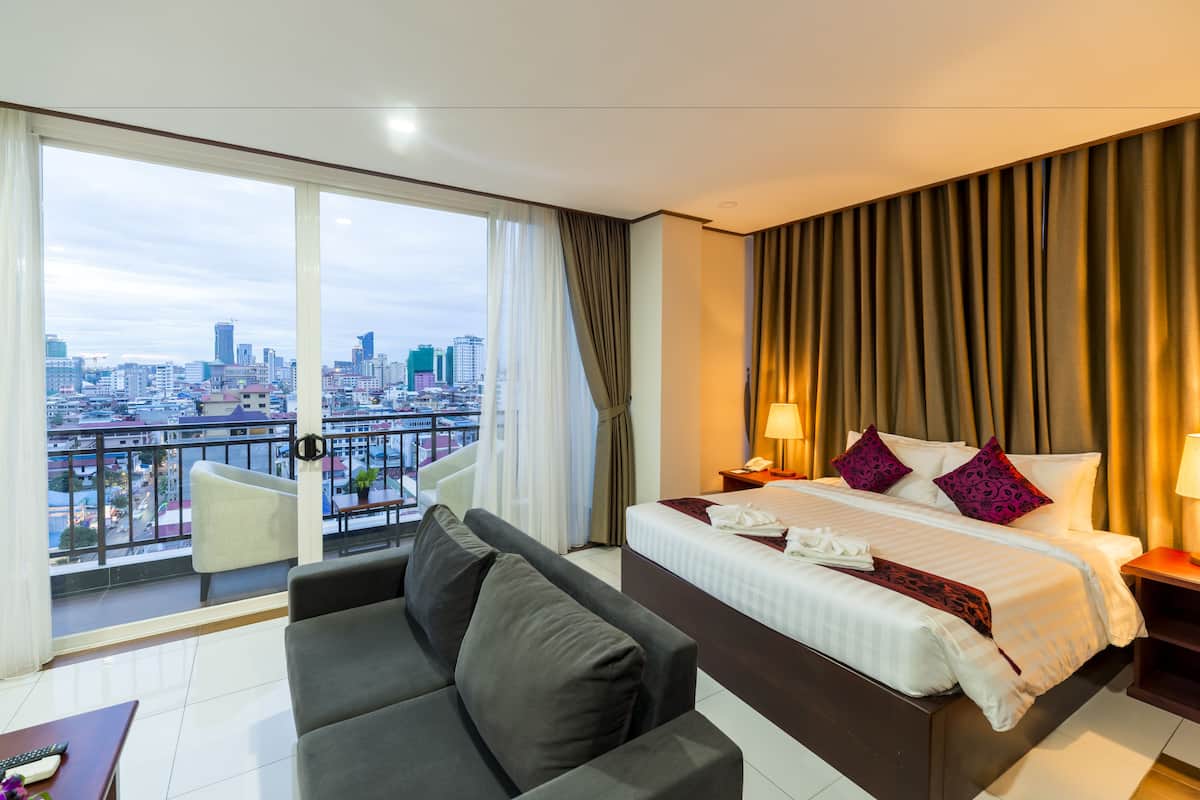 A hotel room with a large bed, a sofa, a balcony with a city view, and a cityscape visible through the windows.