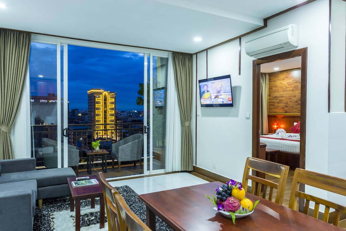A modern hotel room with a balcony, a dining area with a wooden table and chairs, a TV mounted on the wall, and a city view at night.