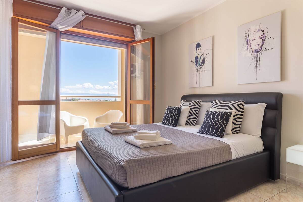 A bedroom with a bed, a view through a sliding glass door, and two artworks on the wall.