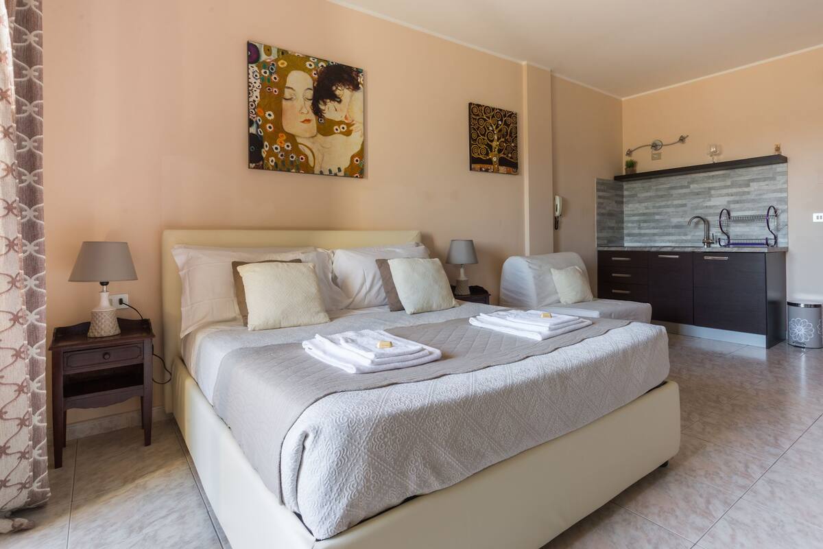 A hotel room with a large bed, bedside tables, a small desk, a mirror, and a kitchenette.
