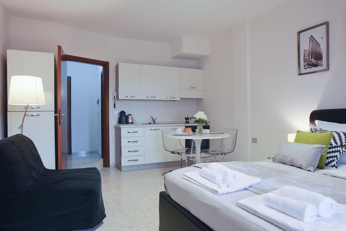 A modern hotel room with a bed, a sofa, a small kitchen area, and a dining table.