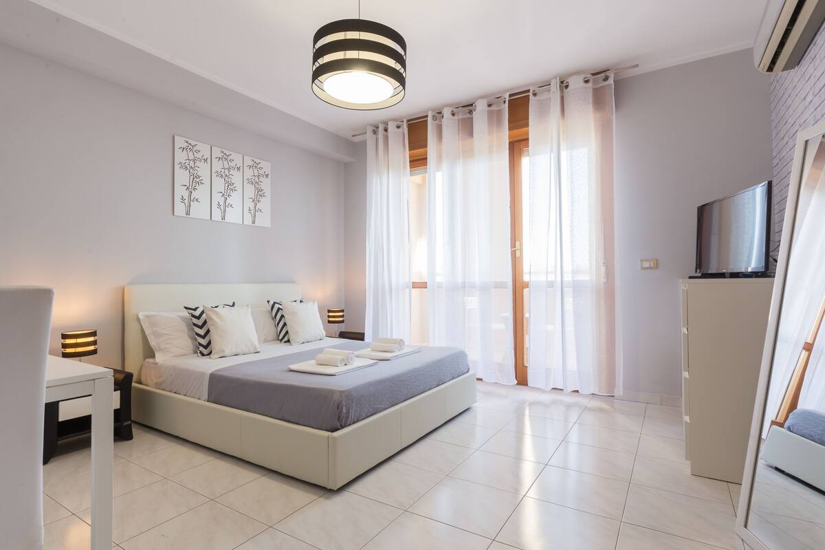 A modern bedroom with a large bed, bedside tables, a flat-screen TV, and a sliding glass door to the balcony.