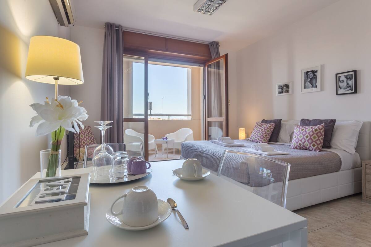A hotel room with a bed, bedside table, lamp, and a view of the outside.