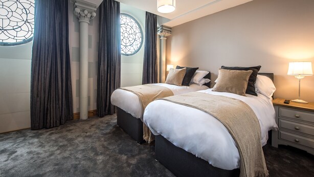 Quarto de hotel com duas camas, uma janela grande com cortinas, um lustre e uma mesa de cabeceira com abajur.
