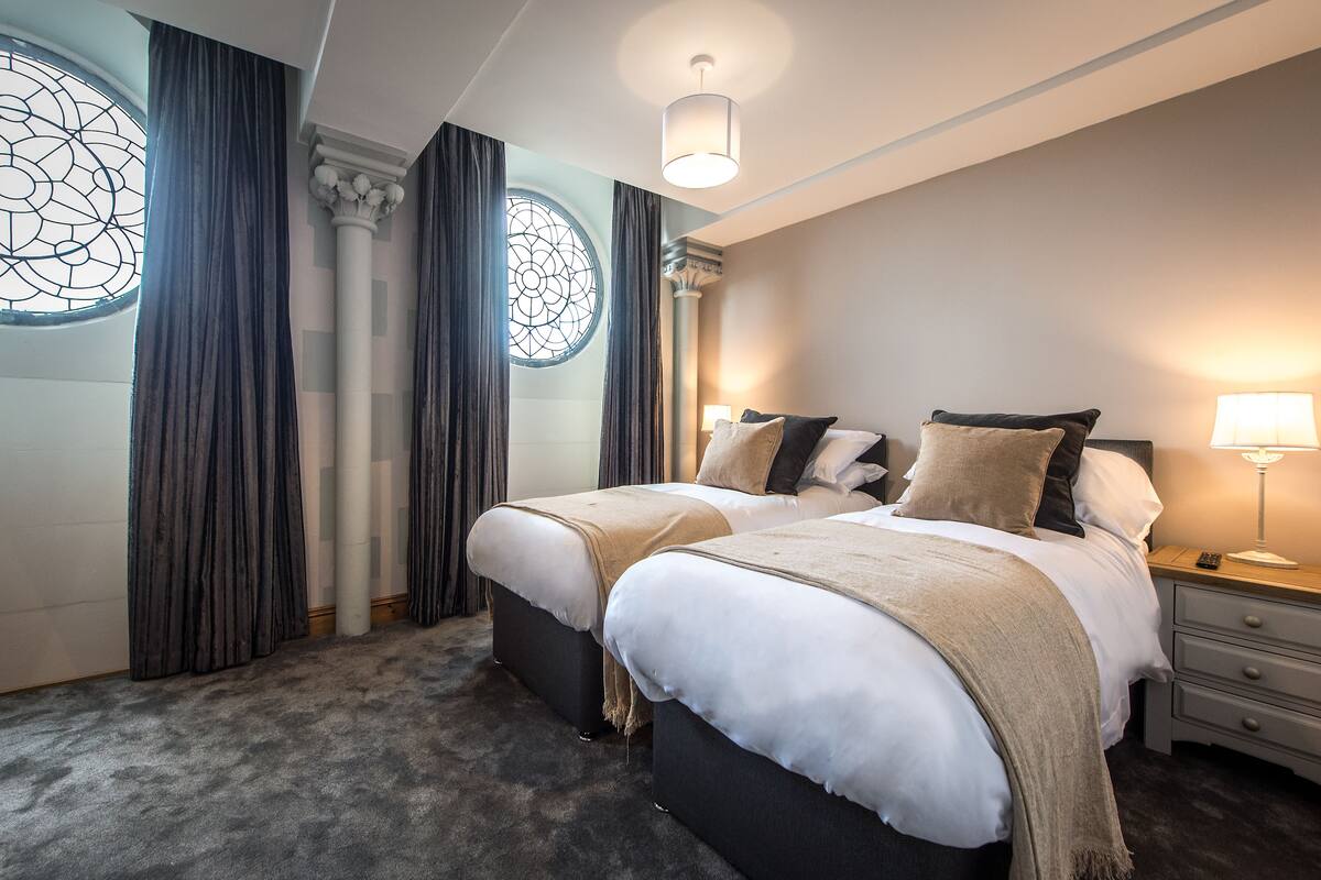 A hotel room with two beds, a large window with curtains, a chandelier, and a bedside table with a lamp.