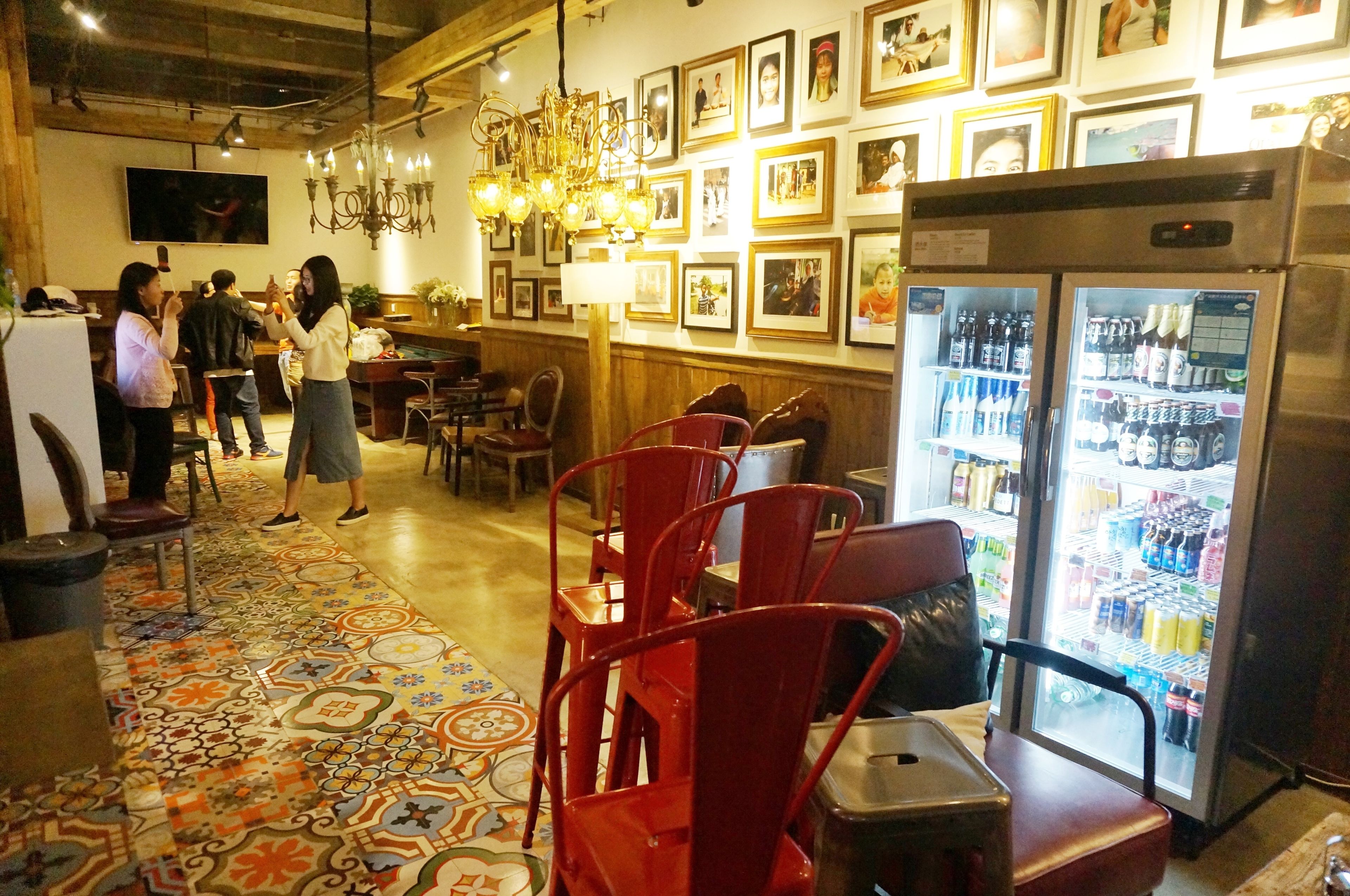 A restaurant interior with a patterned floor, red chairs, a refrigerator with beverages, and framed pictures on the wall.
