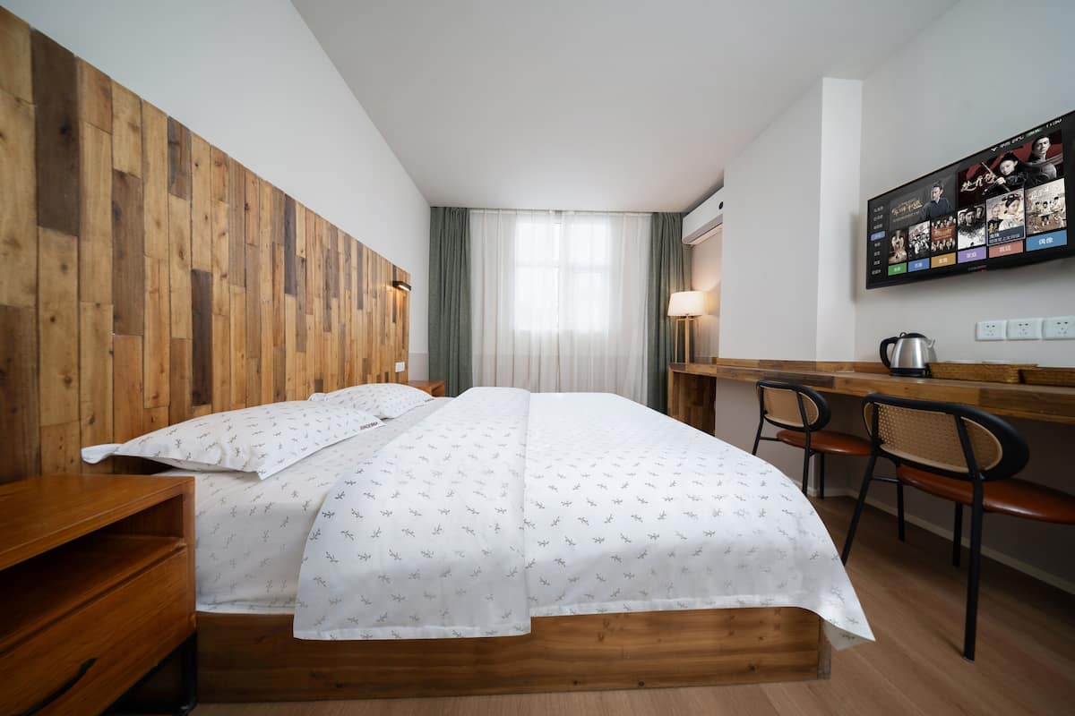 A bedroom with a wooden headboard, a bed with white bedding, a desk with a chair, and a television mounted on the wall.