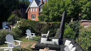 A patio area with outdoor furniture, including a black sofa and white Adirondack chairs, surrounded by greenery and a brick building in the background. - Hôtel du Croisé (Marcq-en-Baroeul)