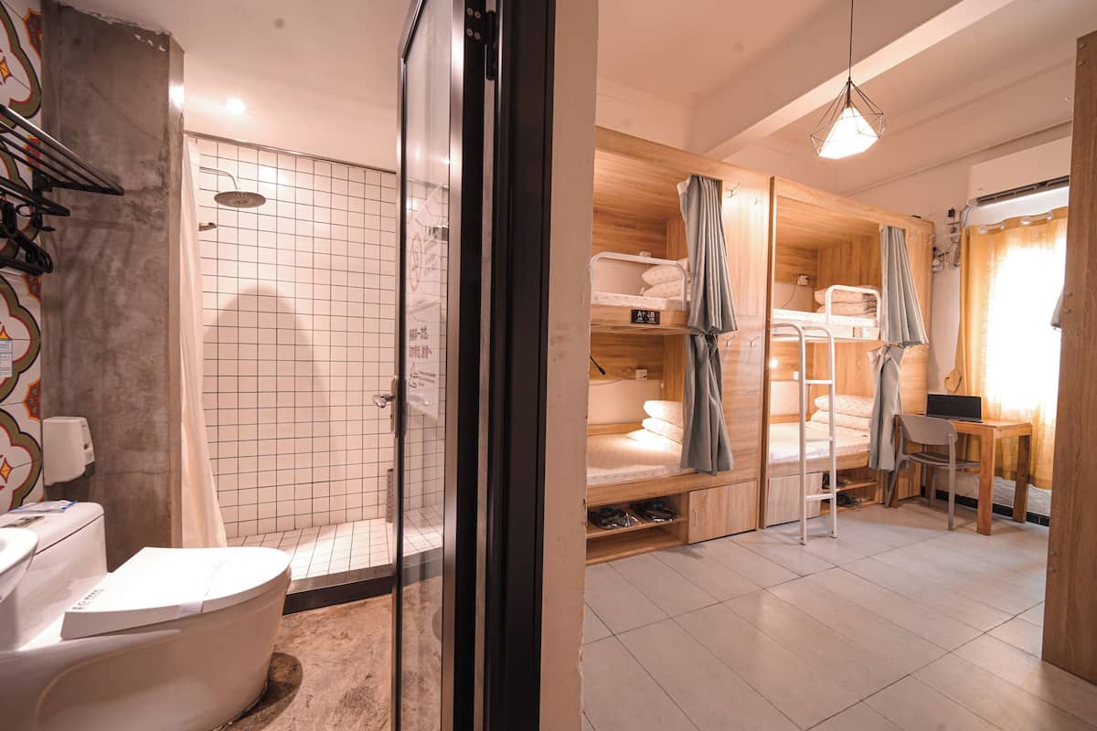 A compact hotel room with a shower, a toilet, a bunk bed, and a small desk with a laptop.