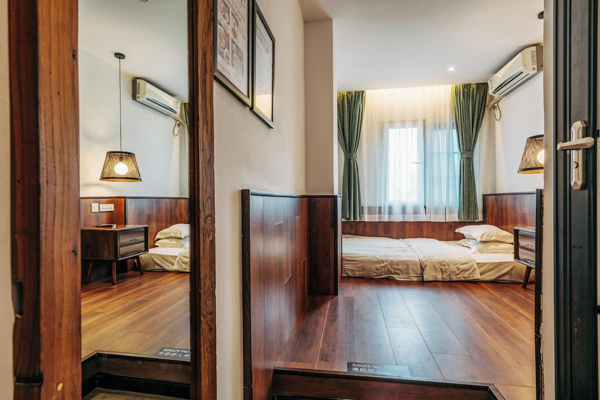 A room with a wooden floor, a bed with a headboard, a bedside table, an air conditioner, and a window with curtains.