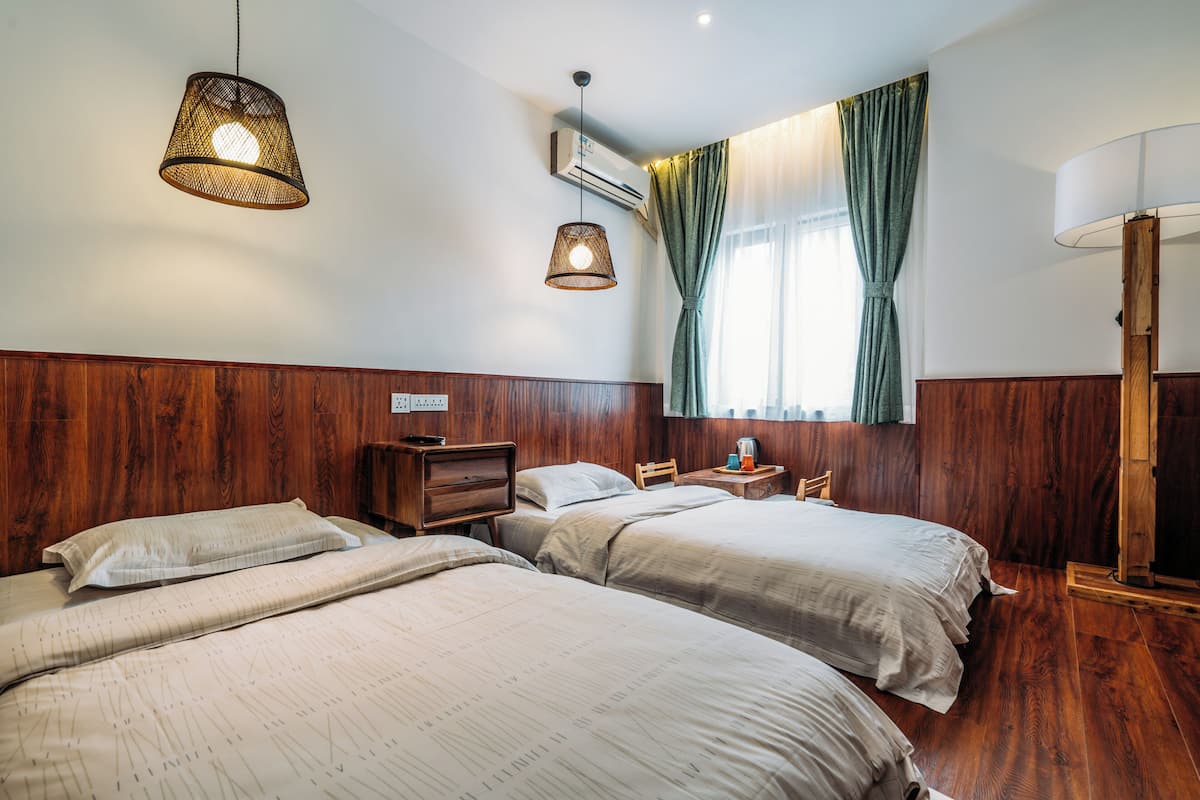 A hotel room with two beds, wooden paneling, a hanging light fixture, an air conditioning unit, and a window with curtains.
