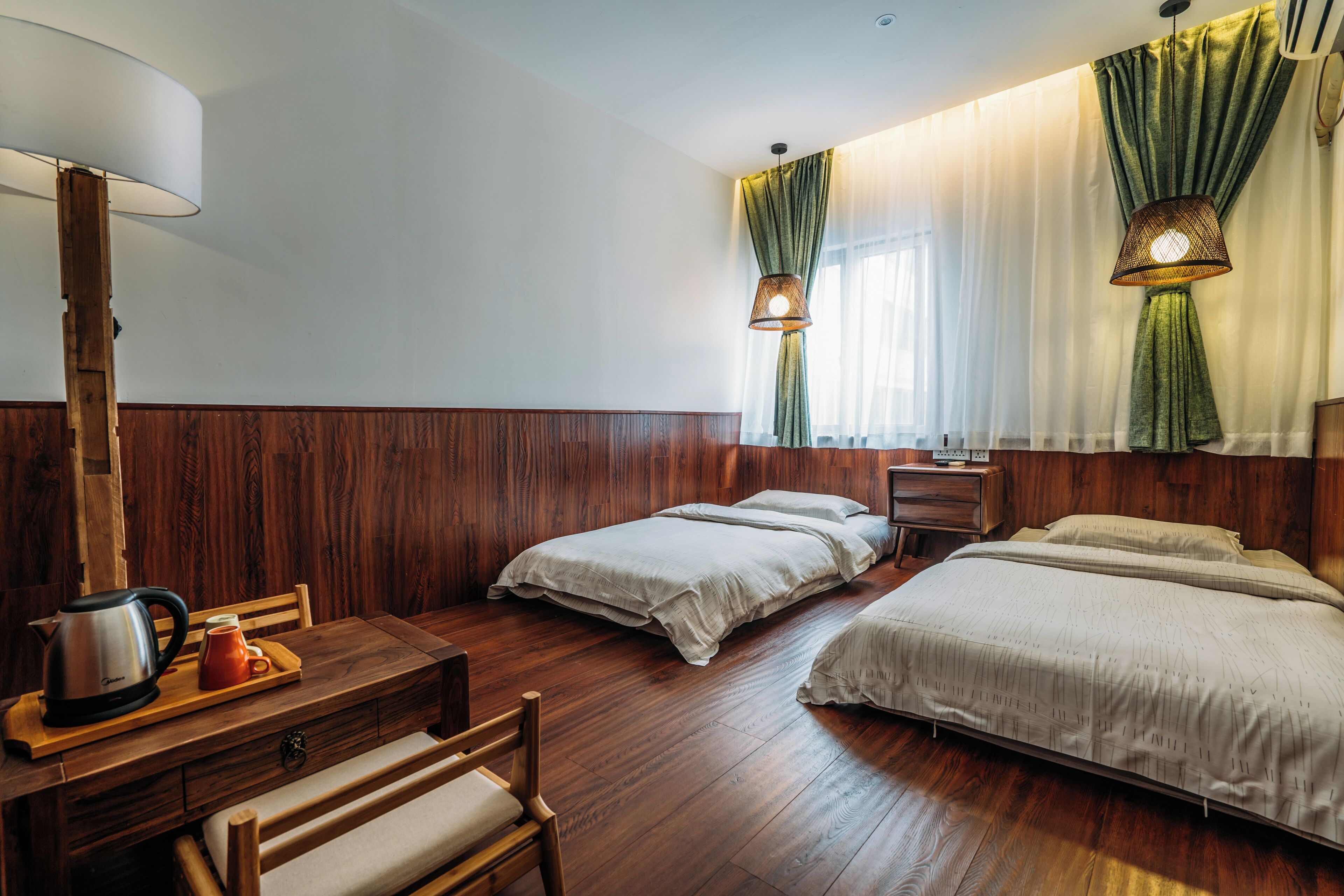 Une chambre d’hôtel avec un plancher en bois, deux lits simples, un bureau en bois avec une bouilloire et une tasse, et une lampe.