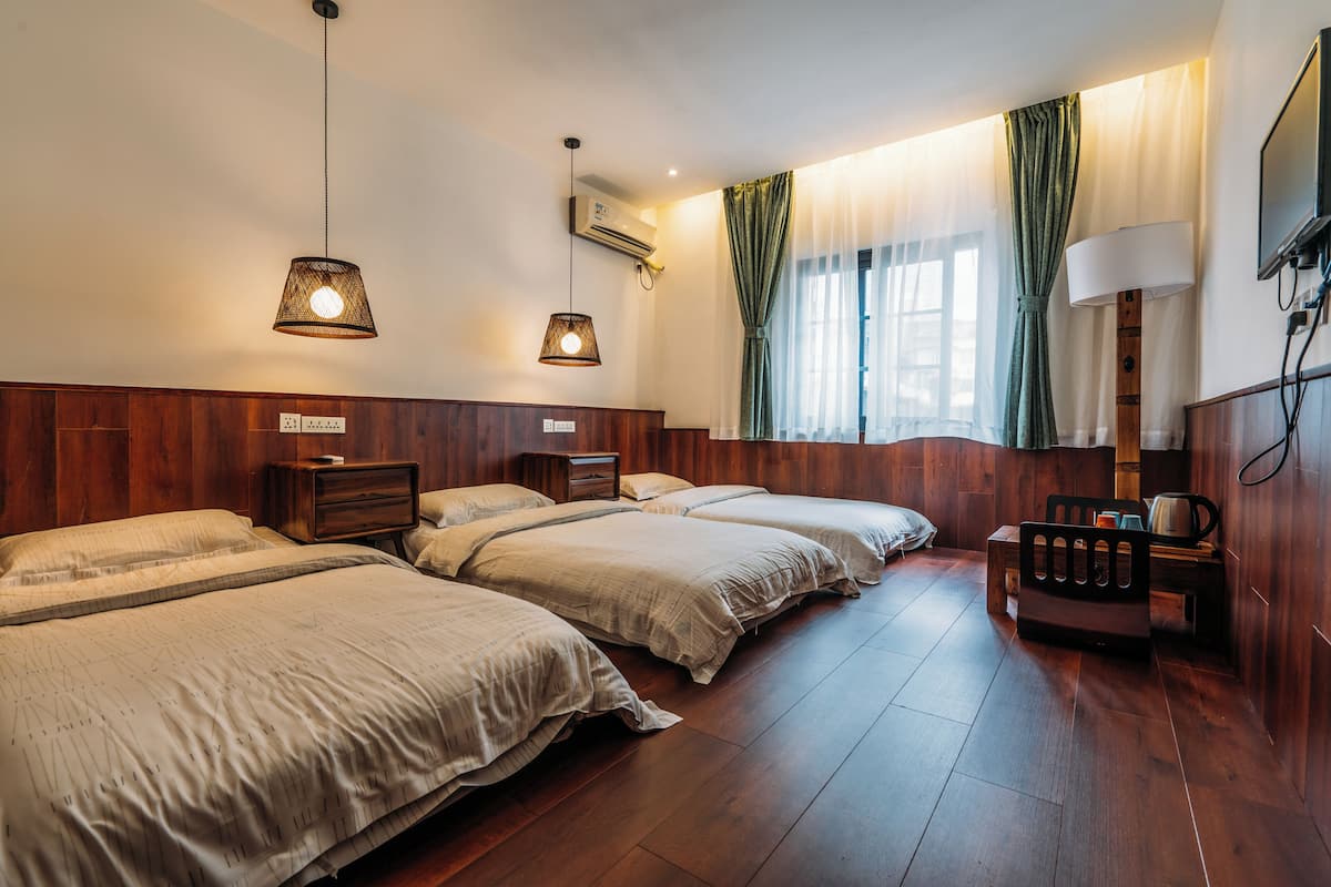 A hotel room with three beds, wooden flooring, a TV, and a bedside table with a lamp.