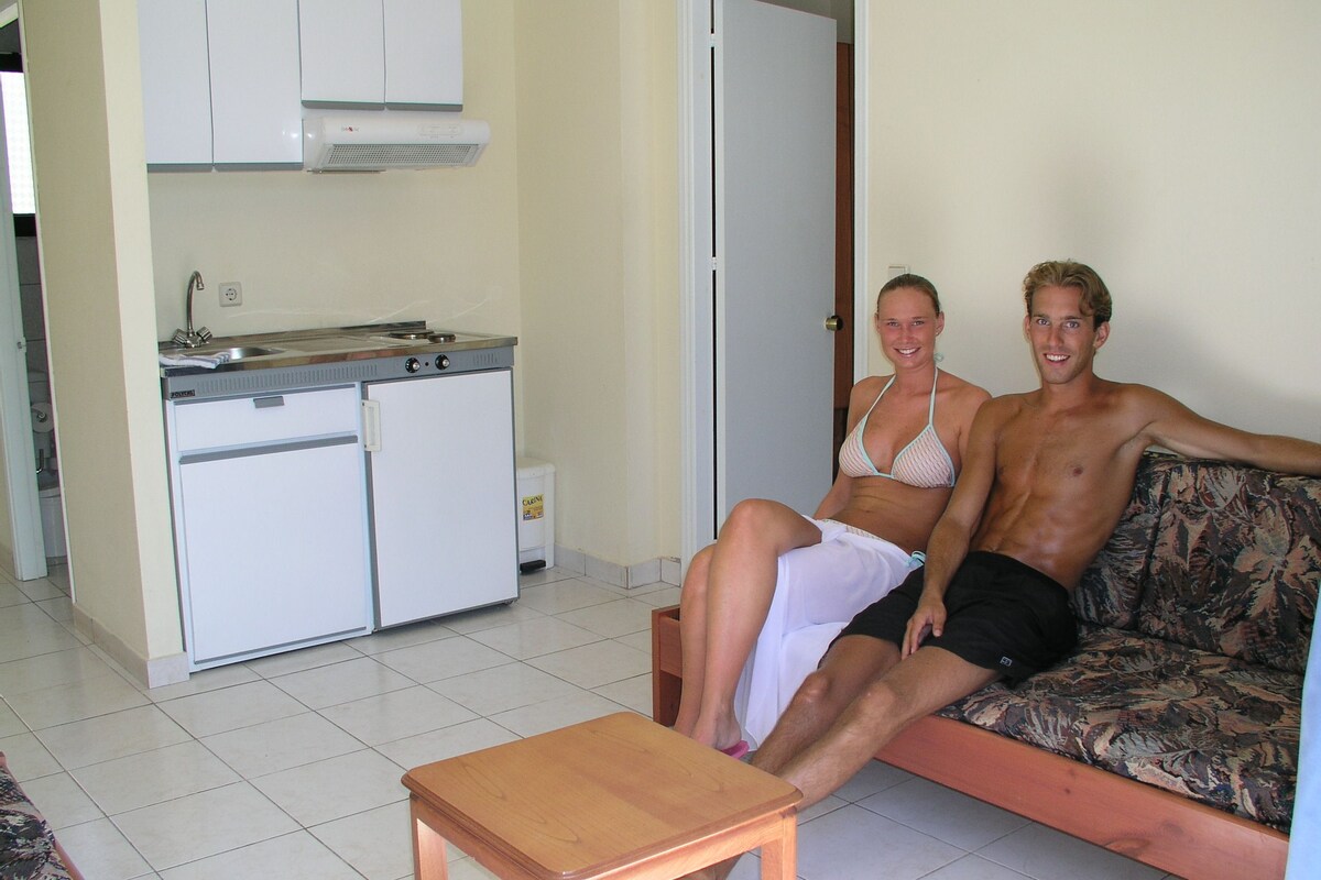 A living room with a sofa, a small wooden table, and a kitchen area with cabinets and a refrigerator.