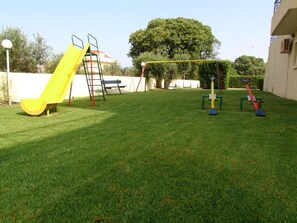 Un espacio al aire libre con césped y equipo de juegos, incluyendo un tobogán amarillo y columpios.