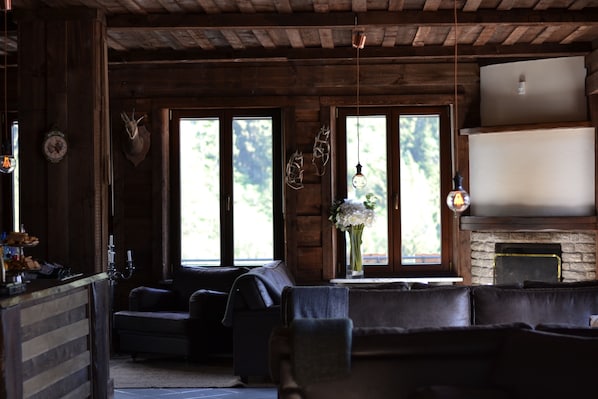 A rustic interior with wooden walls, a stone fireplace, leather sofas, and large windows with a view of greenery.
