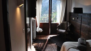 Una habitación con piso de madera, una cama con manta, una silla, un ventanal con cortinas y vistas a montañas.