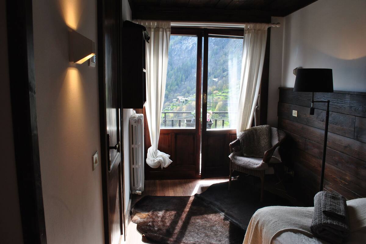 A room with a wooden floor, a bed with a blanket, a chair, a window with curtains, and a view of mountains.