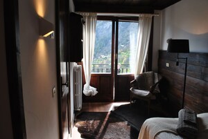 Una habitación con piso de madera, una cama con manta, una silla, un ventanal con cortinas y vistas a montañas.